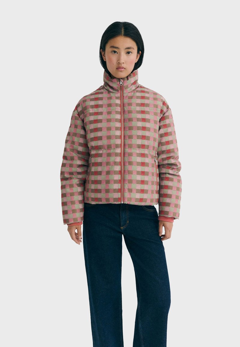 Giacca imbottita in motivo a scacchi rosa e verde, con colletto alto, chiusura a zip e maniche lunghe, abbinata a jeans blu scuro.