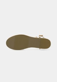 Semelle de sandale à plat sur une surface claire, montrant un dessous texturé beige avec un petit talon et une partie d'une bride blanche avec des clous dorés.