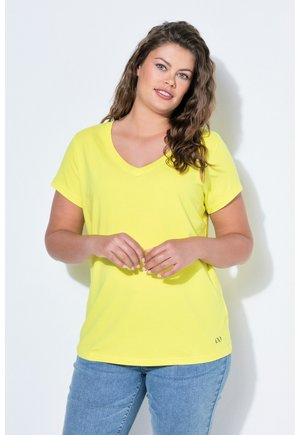 Vrouw met golvend bruin haar die een geel T-shirt met korte mouwen en V-hals en een blauwe spijkerbroek draagt, staand tegen een witte achtergrond.