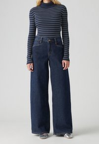 Marinblå randig ribbad polotröja kombinerad med vida, mörka denimjeans. Texturen ser slät ut; jeansen har hög midja och framfickor.
