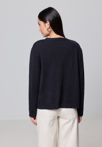 Trampki z granatowego przędzy w luźnym kroju, z okrągłym dekoltem i ściągaczem na dole. Noszone z kremowymi spodniami typu wide-leg. Gładka tekstura.