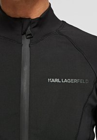 Chaqueta ajustada negra con cremallera, hecha de un tejido suave. Presenta un cuello alto, costuras en contraste y el logo plateado "KARL LAGERFELD" en el pecho.