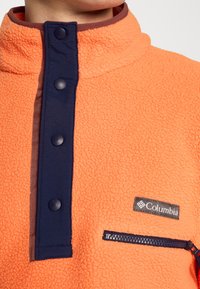 Primer plano de un pull naranja de forro polar con tapeta de botones en azul marino y bolsillo en el pecho con cremallera negra, que presenta un parche con el logo de Columbia.