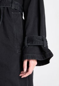 Cappotto in denim nero con maniche lunghe, caratterizzato da una vita con cintura, cuciture a contrasto e tessuto strutturato. Primo piano sul dettaglio della manica.