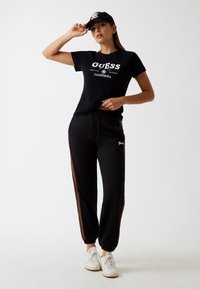 Sort bomulds t-shirt med hvid "GUESS" logo, parret med sorte joggers med brune side-striber og elastiske manchetter, fuldendt med hvide sneakers.