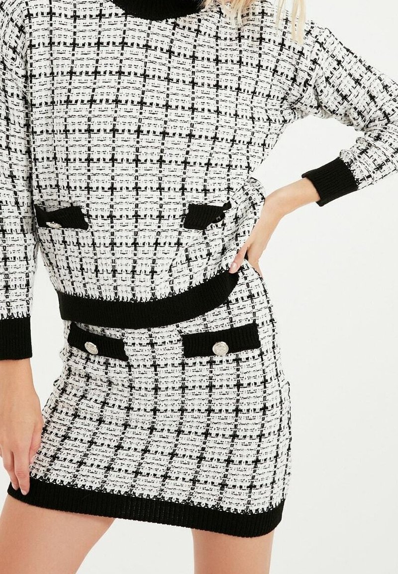 Femme portant un pull tricoté à carreaux noirs et blancs et une jupe assortie avec une bordure noire et des détails de boutons argentés.