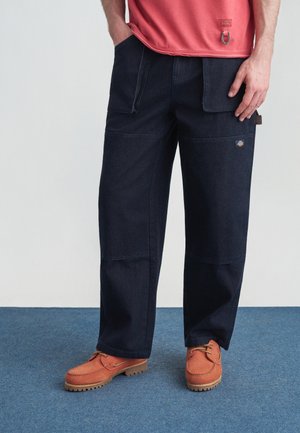 Muž v tmavomodrých pracovných nohaviciach s viacerými vreckami a oranžovo-hnedých šnurovacích topánkach stojaci na modrom koberci.