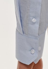 Jasnoniebieski mankiet koszuli z dwoma białymi guzikami, ostrymi krawędziami i teksturowanym materiałem. Mankiet jest starannie złożony z widocznym przeszyciem.