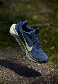 Blå atletiska skor med en ventilerande meshöverdel, gul swoosh-logga, fläckig sula och matchande snören. Höjd över en grusytan.