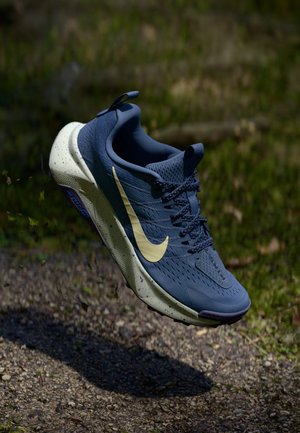 Scarpa sportiva blu con tomaia in mesh traspirante, logo swoosh giallo, suola maculata e lacci coordinati. Elevata sopra una superficie ghiaiosa.