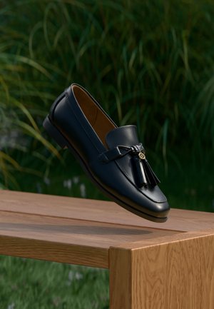 Zwarte leren loafer met kwastjes, rustend op de rand van een houten oppervlak, met groene bladeren vaag op de achtergrond.