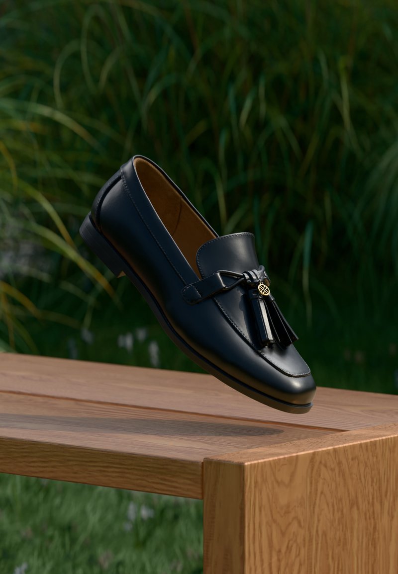 Μαύρο δερμάτινο μοκασίνι με φούντες που ακουμπά στην άκρη μιας ξύλινης επιφάνειας, με πράσινη φυλλωσιά θολωμένη στο φόντο.