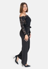 Body in pizzo nero con maniche scoperte, abbinato a pantaloni neri a vita alta. La modella indossa tacchi a punta neri. Texture liscia.