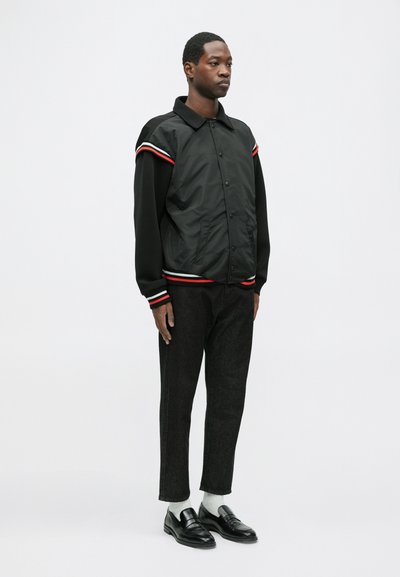 Mustriline must jakk punaste ja valgete triipudega õlgadel, nööbiga sulgemine ja kootud varrukad, koos tummiste pükste ja mustade loaferitega.