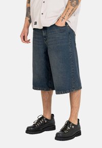 Dunkelblaue Jeansshorts, lockerer Schnitt, bis zur Mitte des Knies reichend, mit genähtem Saum. Kombiniert mit schwarzen Schnürschuhen mit metallischen Akzenten.