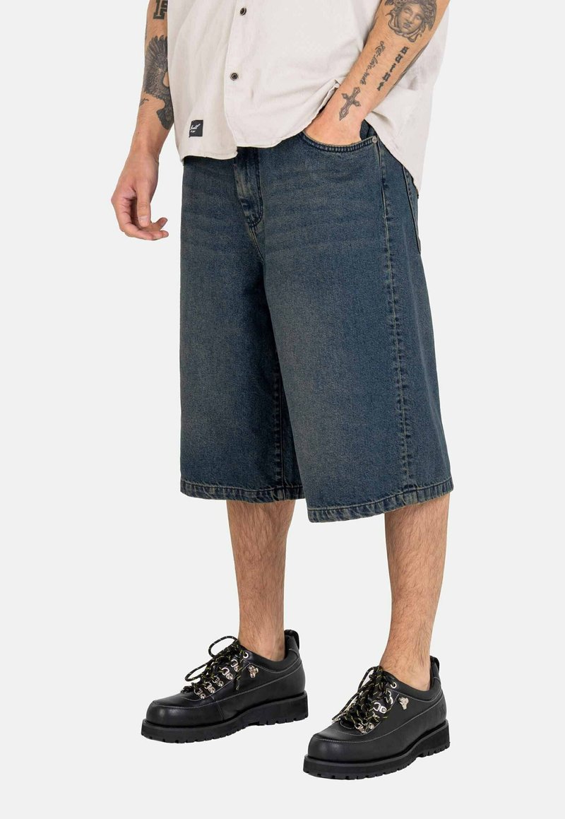 Dunkelblaue Jeansshorts, lockerer Schnitt, bis zur Mitte des Knies reichend, mit genähtem Saum. Kombiniert mit schwarzen Schnürschuhen mit metallischen Akzenten.