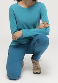 Turquoise long-sleeve shirt with a textured fabric, paired with matching blue pants and tan hiking boots. Hands resting on knees.