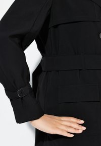 Schwarzer Trenchcoat mit tailliertem Gürtel, ausgestattet mit einem geknöpften Ärmelbund und einer Klappentasche an der Seite, aus glattem Stoff gefertigt.