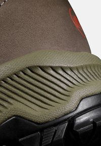 Bruine suede laars met een getextureerde olijfgroene rubberen zool, voorzien van ribbels en een zwarte gripzool, en rode logodetails.