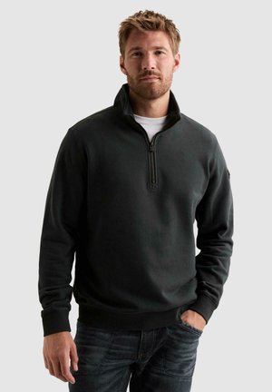 Homme aux cheveux blonds courts et barbe, portant un pull noir à fermeture éclair quart de zip sur une chemise blanche, jean bleu, debout avec une main dans la poche.
