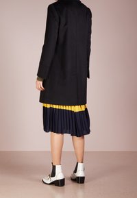 Cappotto di lana nera, gonna plissettata gialla e blu navy, stivaletti bianchi alla caviglia con dettagli neri e un sottogiacca a maglia strutturata. Design minimalista.