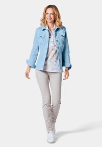 Lichtblauwe spijkerjack met twee borstzakken, over een bloempatronen shirt. Lichtbeige broek en witte sneakers maken de outfit compleet.