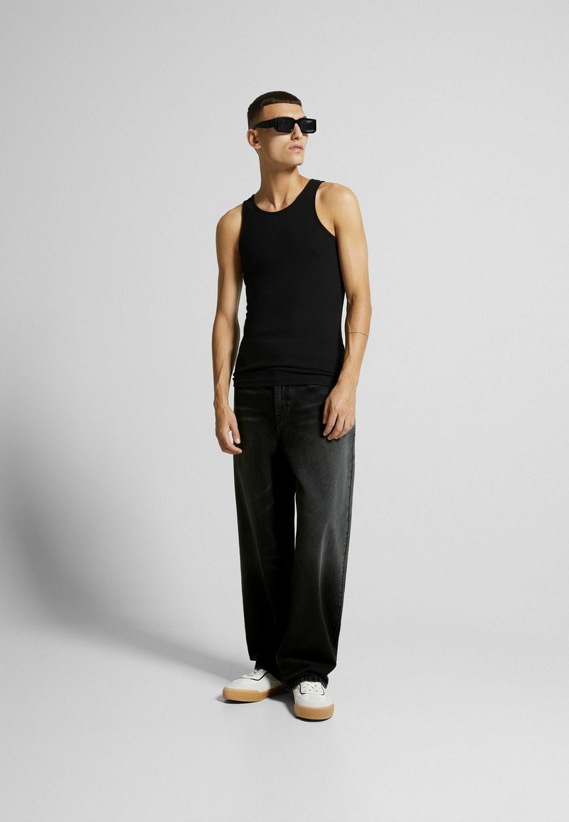 Top nero senza maniche, jeans larghi in denim scuro, sneaker bianche con suola in gomma e occhiali da sole neri a forma quadrata. Sfondo semplice.