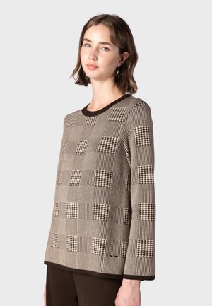Jeune femme portant un haut à manches longues beige et marron à carreaux avec une bordure marron foncé, debout devant un fond clair uni.