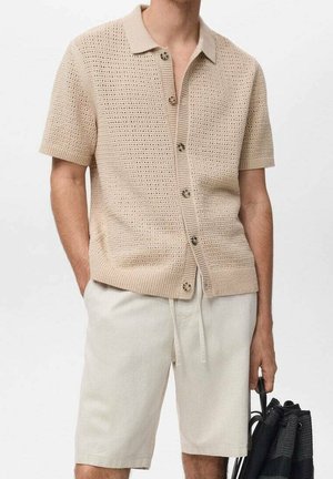 Mann trägt beige kurzärmliges Strickhemd mit Knöpfen und hellbeige Shorts mit Kordelzug, hält eine schwarze geflochtene Tasche in der rechten Hand.
