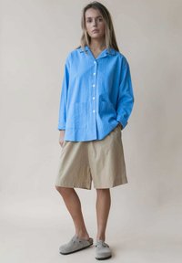 Lichtblauw overhemd met kraag, twee voorkeurszakken, gecombineerd met beige knielange shorts en grijze instappers. Glad textuur.