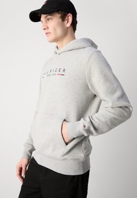 Svijetlosiva hoodie s prednjim džepom koja sadrži logo "HIL FIGER NEW YORK" u tamnoplavoj i crvenoj boji. Rukavi su manžetasti s malim akcentom loga.
