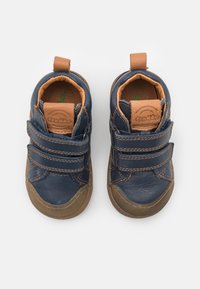 Chaussures en cuir bleu marine avec des accents marron, dotées de deux bandes Velcro, d'un bout rond et d'une semelle en caoutchouc beige. Doublure intérieure douce visible.