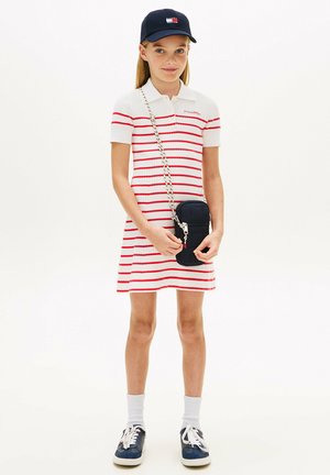 Meisje draagt een wit met rood gestreepte jurk, een marineblauw petje, witte sokken, marineblauwe sneakers, en houdt een kleine marineblauwe crossbodytas vast.