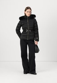 Black puffer jacket with a fur-trimmed hood, zipped front, belted waist, and logo detail. Paired with wide-leg black jeans and a small handbag.