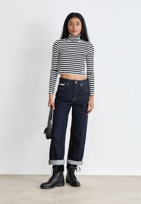 Woman wearing black and white striped turtleneck crop top, dark cuffed jeans, black lace-up boots, holding small black handbag, standing on plain floor.