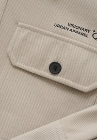 Beige shirt van textiel met textuur, voorzien van een klepzak, zwarte knoop en de opdruk "VISIONARY URBAN APPAREL." Het shirt heeft een diagonaal ribbelpatroon.