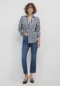 Camicia button-up a righe blu navy e bianche, abbinata a jeans cropped blu scuro e tacchi a punta neri. La camicia ha le maniche arrotolate.
