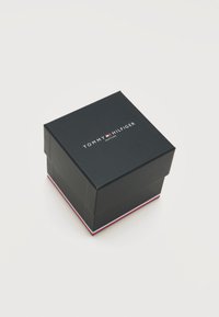 Μαύρο κουτί ρολογιού Tommy Hilfiger με το λογότυπο και κόκκινες, λευκές και μπλε ρίγες γύρω από τη βάση, σε λευκό φόντο.