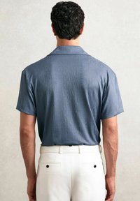 Cămașă cu mânecă scurtă dintr-un material texturat albastru, cu guler, având un model de dungi verticale, combinată cu pantaloni albi și un detaliu cu nasturi negri.