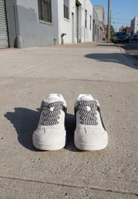 Ein Paar weiße Sneakers mit schwarzen Zickzack-Schnürsenkeln, die auf einem Bürgersteig in der Stadt vor einer Reihe von Gebäuden und geparkten Autos stehen.