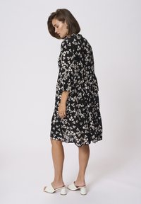Robe noire à fleurs avec des manches courtes, un design évasé et des motifs de fleurs roses pâles. Portée avec des mules blanches à talons.