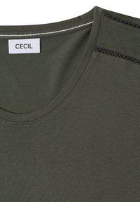 T-shirt col rond vert olive avec étiquette de marque blanche "CECIL" cousue à l'intérieur du col et détails de coutures visibles sur les épaules.