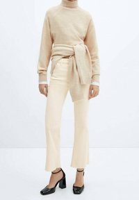 Pull à col roulé beige, détail de ceinture à la taille ; associé à un jean clair évasé avec un ourlet non fini ; chaussures à talons carrés noires avec des brides au niveau de la cheville.