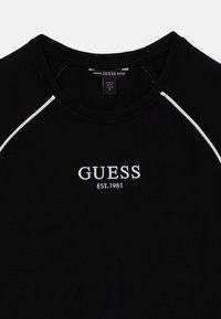 Fekete póló kerek nyakkivágással és rövid ujjakkal. Fehér szegéllyel díszített a varrások mentén, valamint a "GUESS EST. 1981" fehér hímzett logóval.