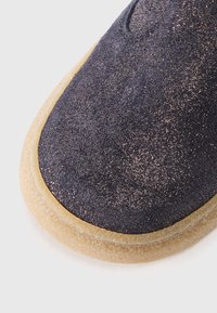 Pantoufle en daim bleu foncé avec une finition texturée scintillante, bout rond et semelle en caoutchouc de couleur claire. Présente un design simple à enfiler.