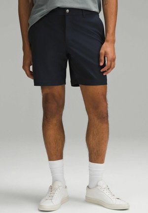 Pantalones cortos azul marino de tejido ligero, con un diseño de frente plano, bolsillos y cierre de botón. Se usan con zapatillas blancas y calcetines hasta la altura del tobillo.