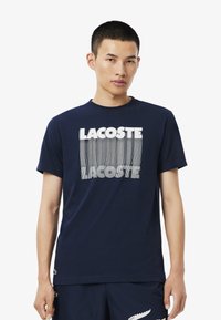 Camiseta de algodón azul marino con cuello redondo y mangas cortas, que presenta un gráfico geométrico blanco del logo "LACOSTE" en la parte frontal.