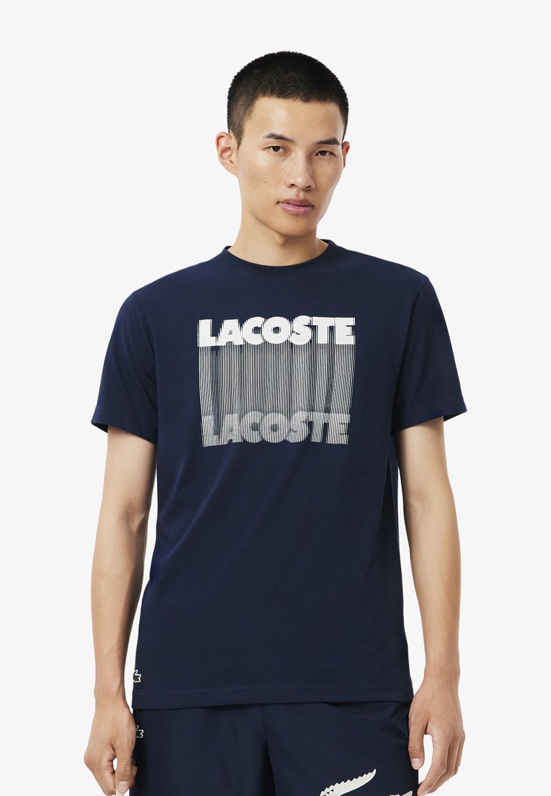 Camiseta de algodón azul marino con cuello redondo y mangas cortas, que presenta un gráfico geométrico blanco del logo "LACOSTE" en la parte frontal.