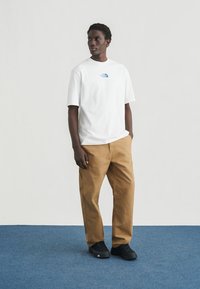 Λευκό βαμβακερό t-shirt με μπλε λογότυπο, συνδυασμένο με καφέ παντελόνι και μαύρα αθλητικά παπούτσια. Το μοντέλο στέκεται μπροστά σε ανοιχτό γκρι φόντο.