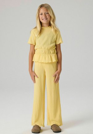 Joven chica de pie y sonriendo, con una camiseta amarilla texturizada de manga corta con detalle de volantes y pantalones anchos a juego con zapatos marrones.